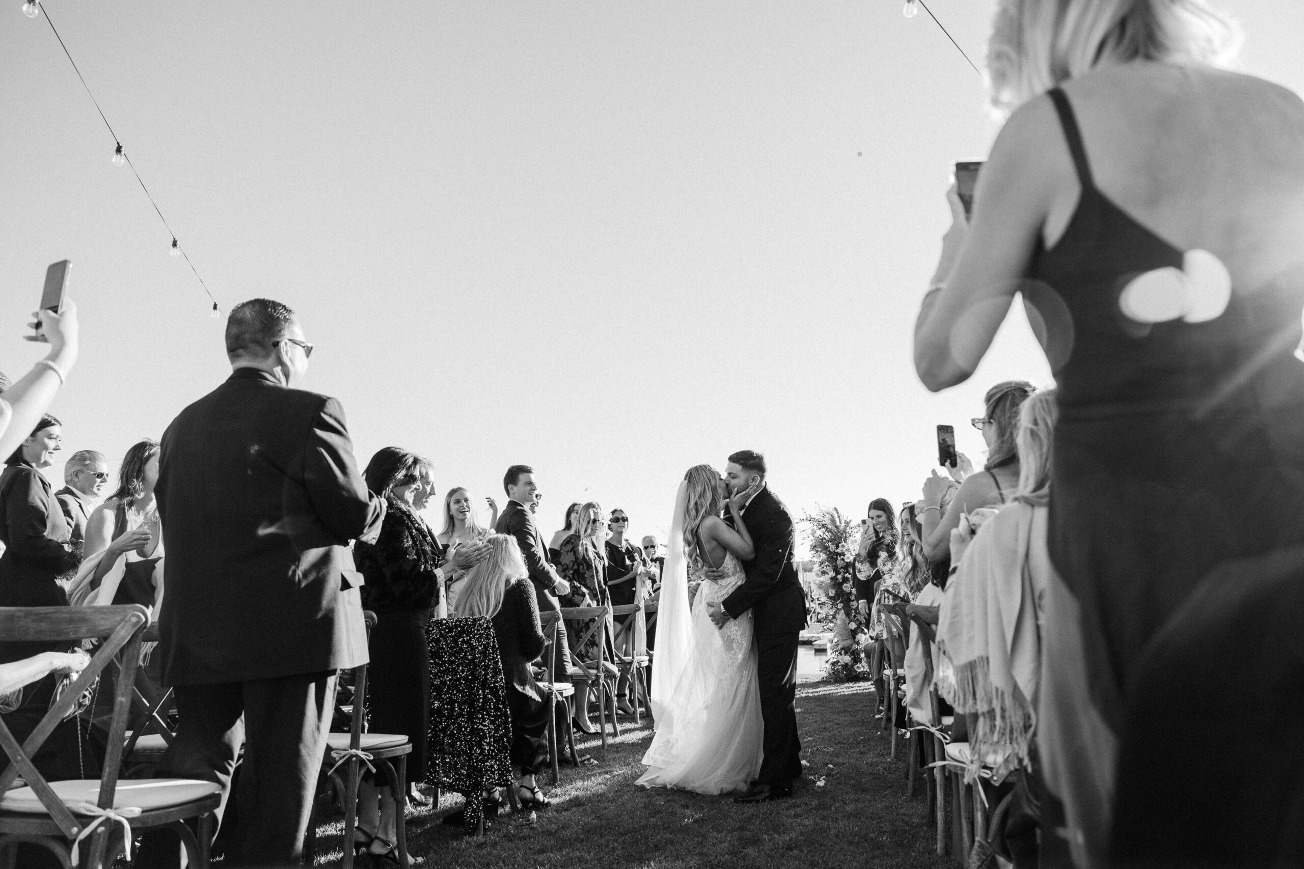 black and white photograph of newly married couple kissing in the aisle during recessional as guests applaud them.