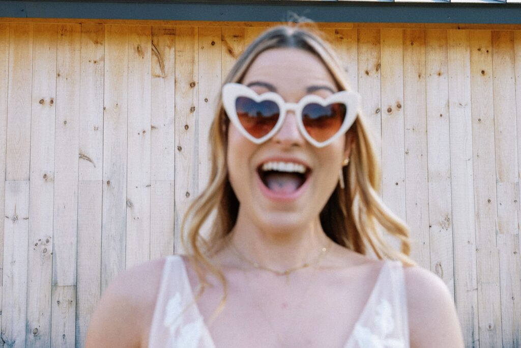 a bride smiles wide while wearing heart shaped sunglasses, she is out of focus with the wood fence behind her in focus