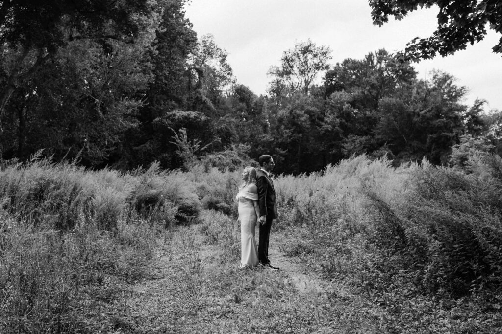 a pair of newlyweds stand back to back in an empty field during photos with their wedding photographer