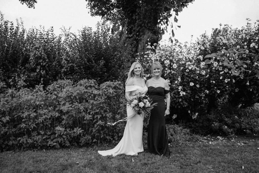 a woman wearing a wedding dress and holding a bouquet stands smiling next to her mother during photos with a wedding photographer