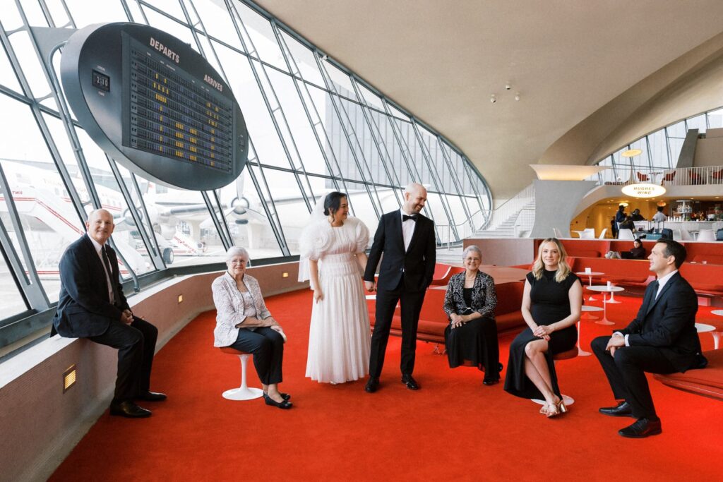 a pair of newlyweds and their close relatives sit in an airport smiling at each other during photos with their wedding photographer at the TWA Hotel