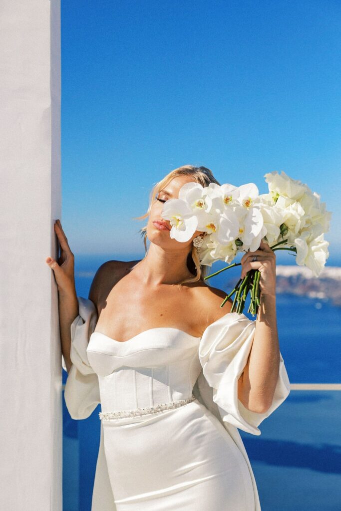 a woman wearing a white dress leans against a white building with the blue sky and sea behind her holding a bouquet in front of her face during photos with a wedding photographer in Santorini Greece