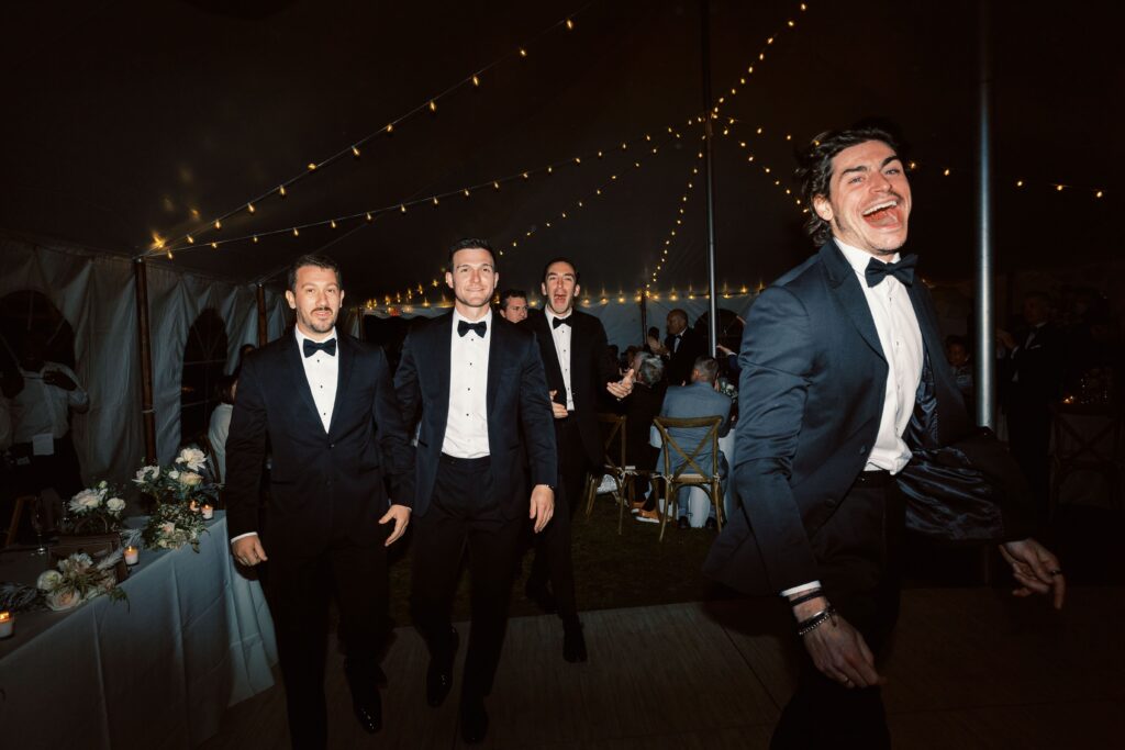 a handful of men with excited expressions smile in tuxes as they walk by the wedding photographer