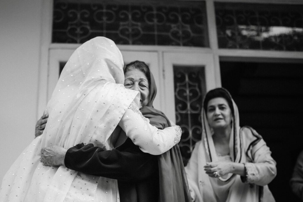 a woman wearing white hugs her grandmother with her mother standing behind them facing the wedding photogrpaher