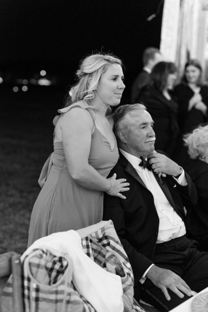 a daughter stands behind her seated father during an emotional speech from the groom at a wedding at kontokosta winery in long island.