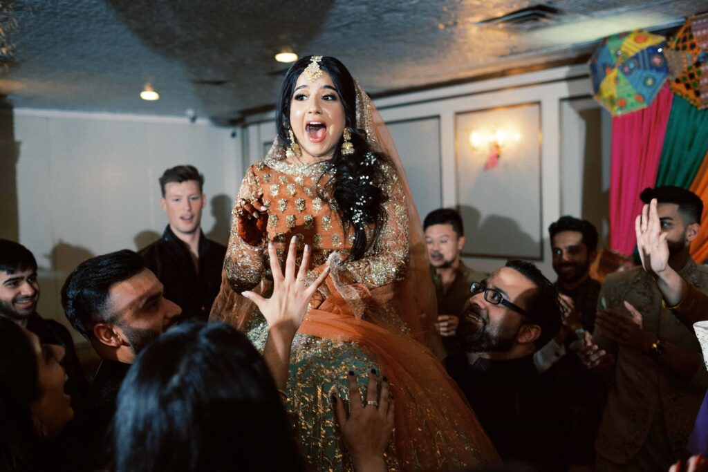 a woman, open mouthed in surprise, is lifted on a group of people's shoulders at a south asian wedding