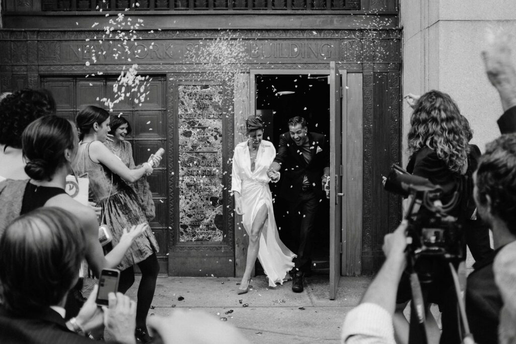 wedding photographer captures newlyweds leaving a new york city hall building beaming as their friends and family throw rice and confetti at them